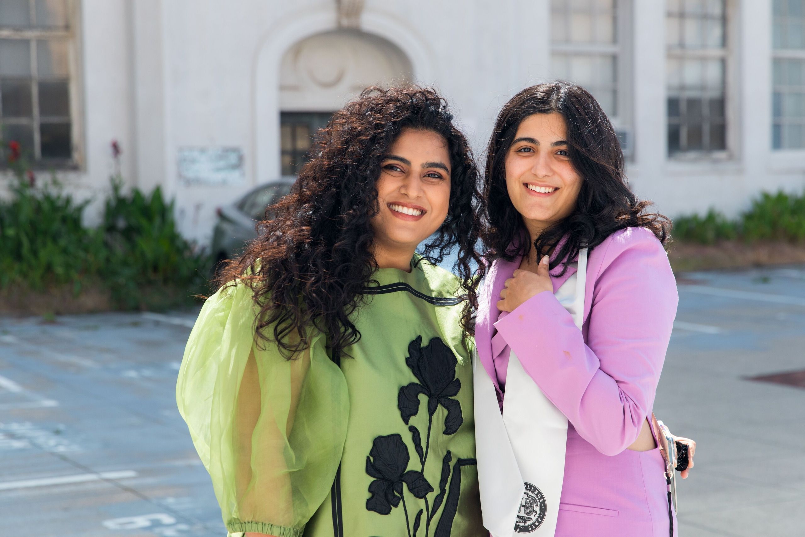 Two graduates in formal attire pose together outdoors; one wears a green dress and the other a pink blazer, both smiling in front of a white building.
