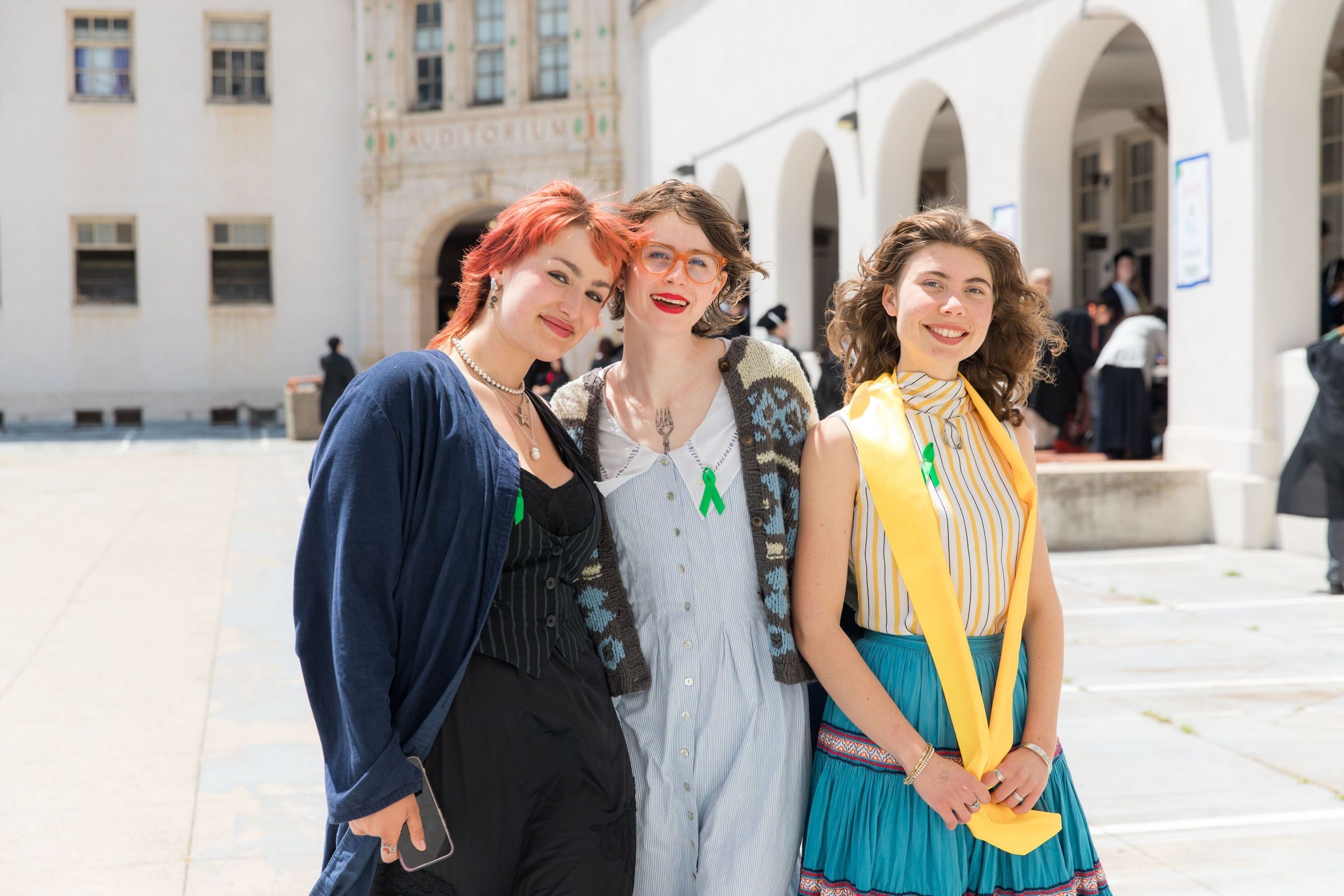 Three graduates with colorful hair stand together on a white courtyard; one wears navy and black, another a gray dress, and the third a teal skirt with yellow sash.