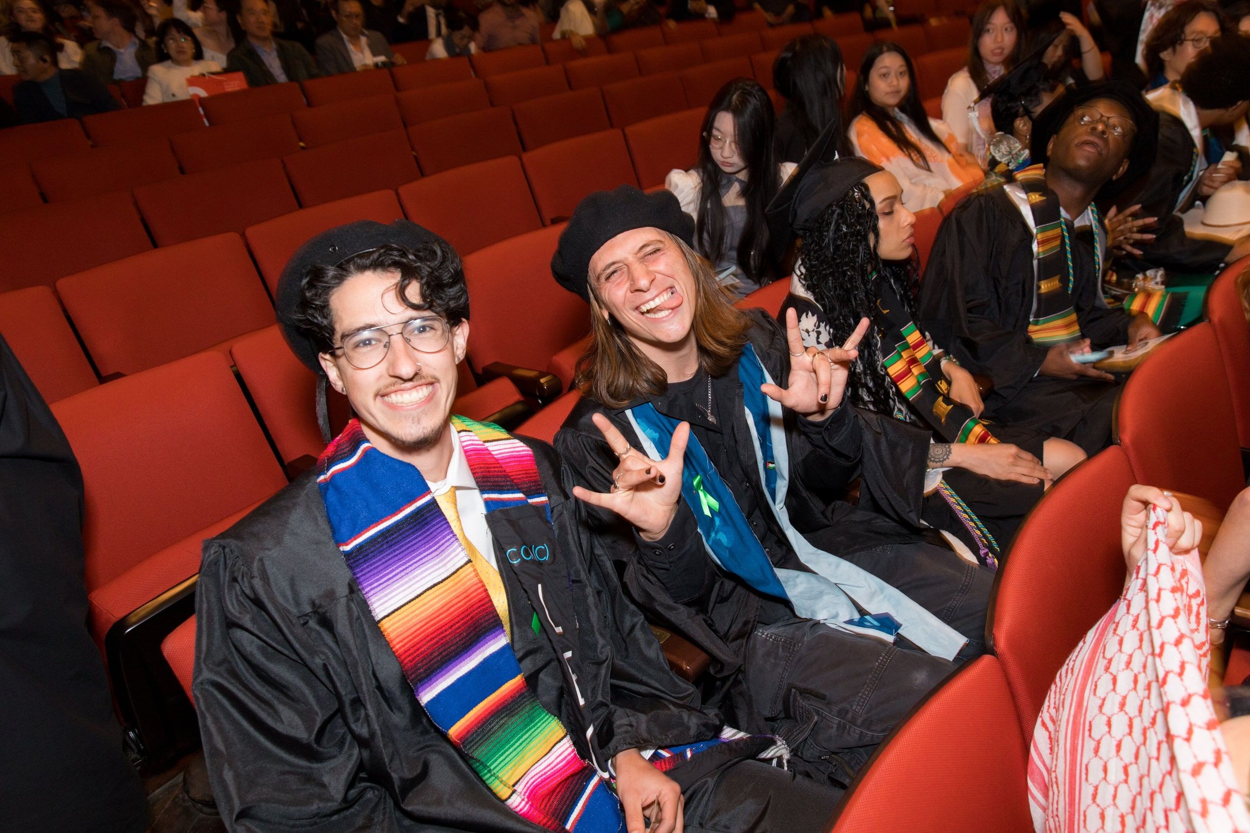 Two graduates in black robes with colorful cultural sashes sit together on red auditorium seats, smiling during the commencement ceremony.