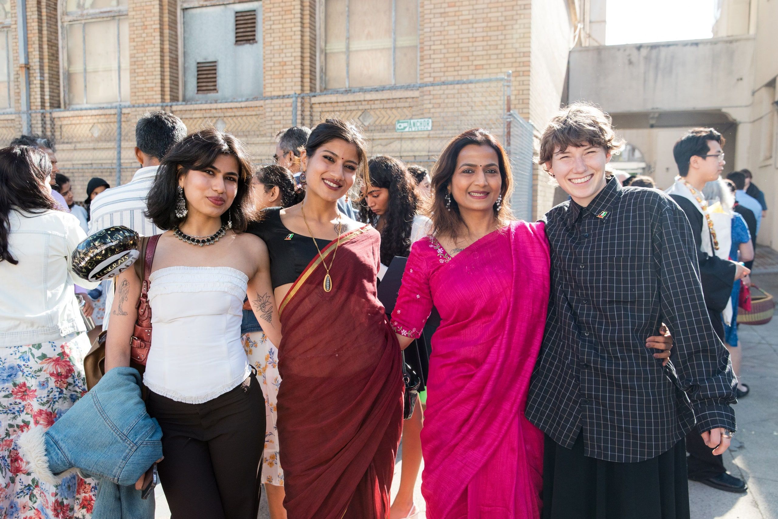 Four graduates pose together outdoors against a brick building; they wear a mix of casual and formal attire, including vibrant pink and maroon colors.