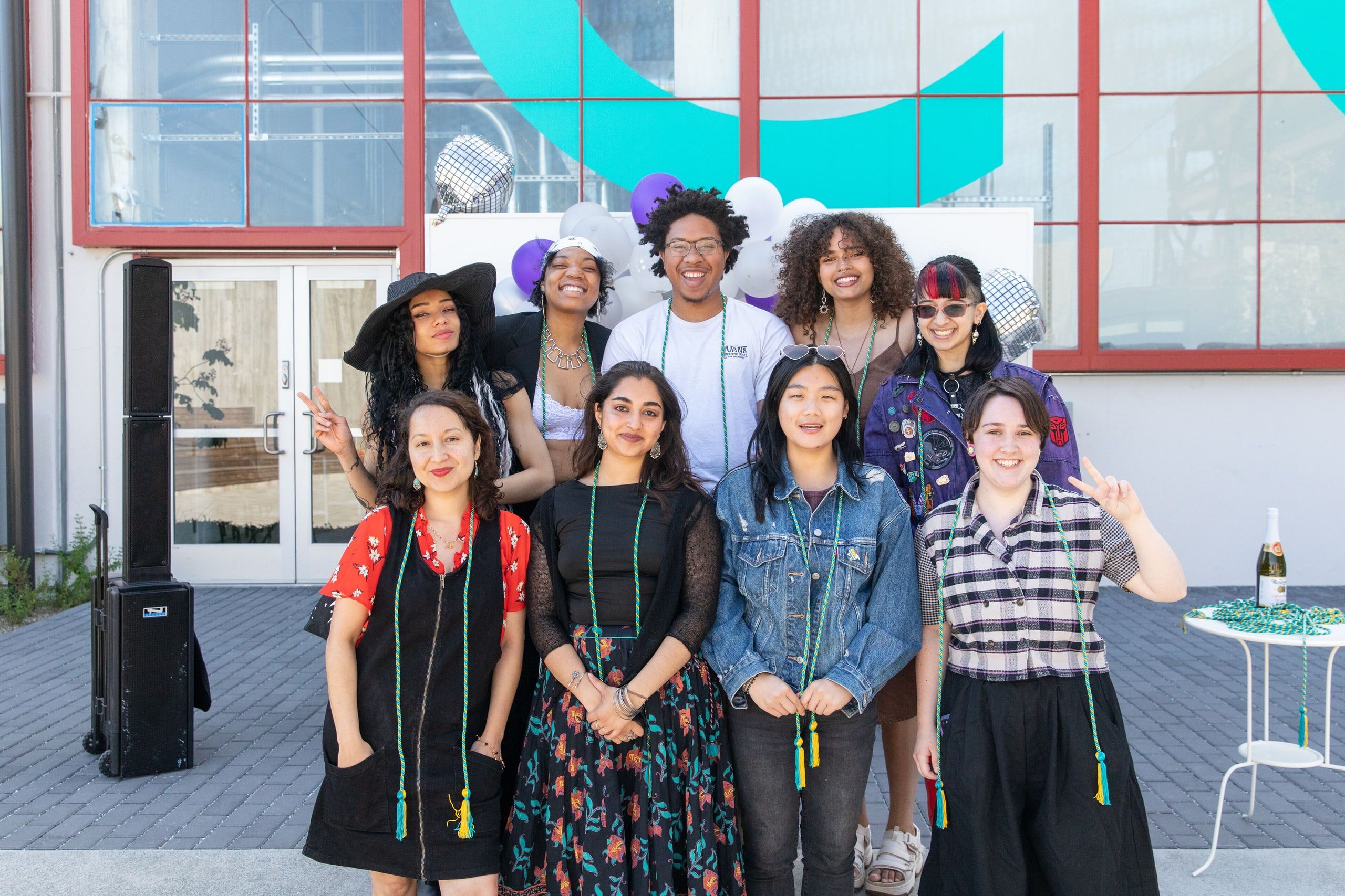 A diverse group of young graduates wearing leis and graduation regalia pose together outdoors in front of a modern building with colorful turquoise architectural elements.
