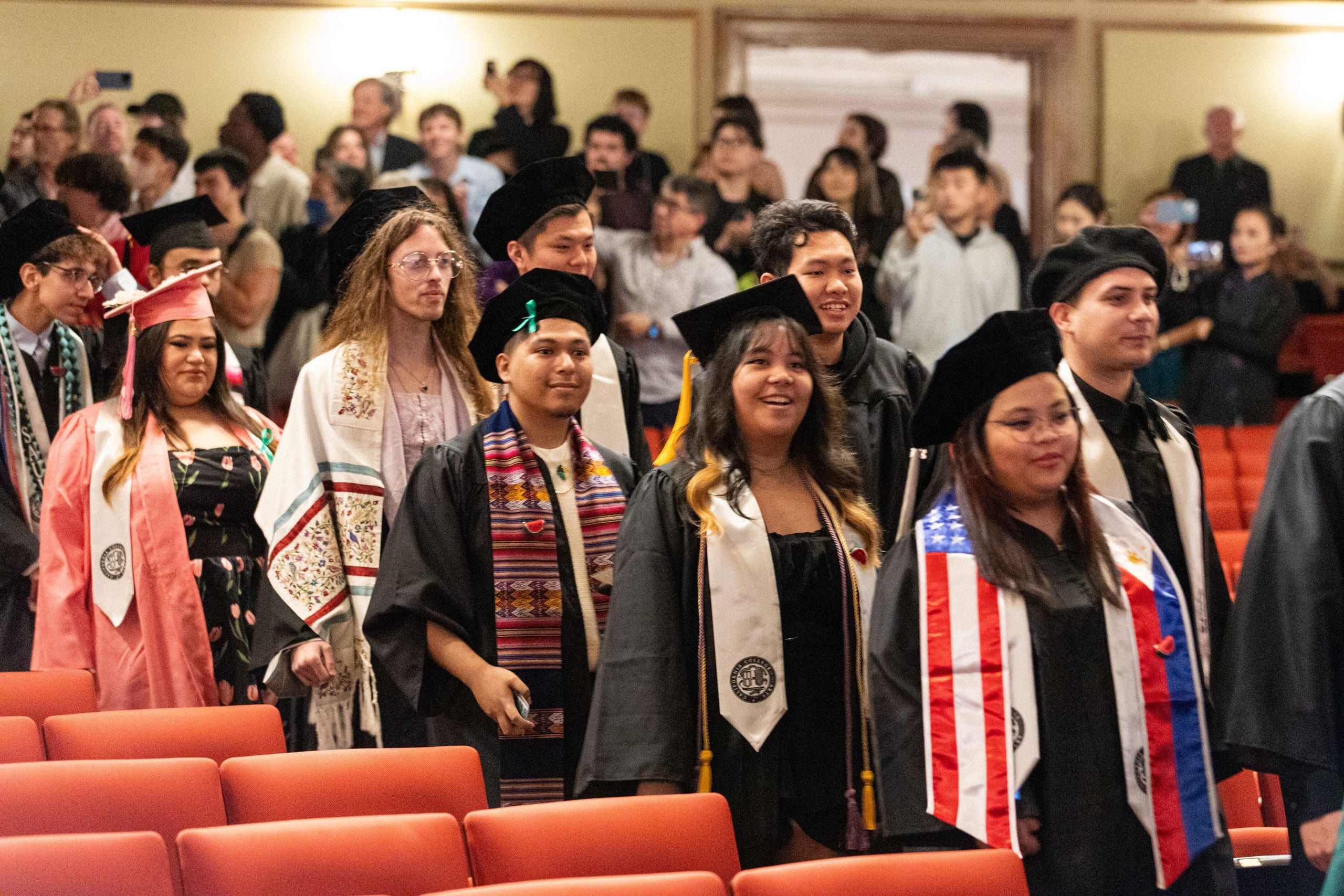 Graduates in caps, gowns, and cultural regalia stand in an auditorium with red seats, many wearing decorative sashes and stoles representing their cultural heritage.