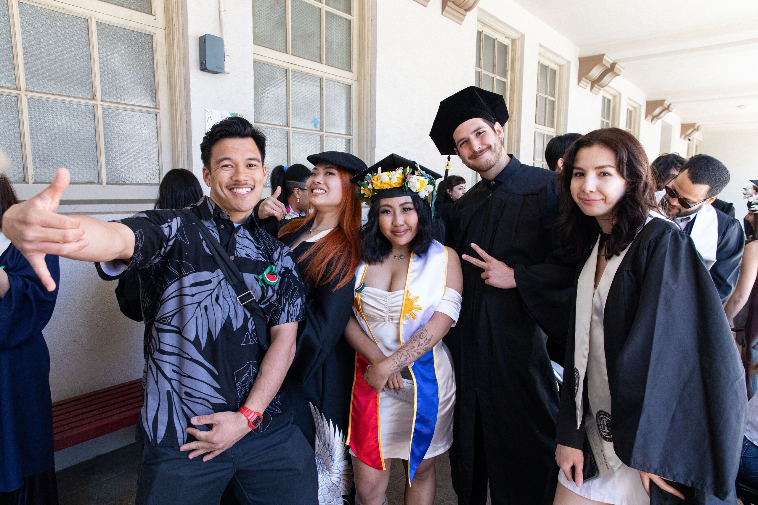 A group of graduates in caps and gowns pose together indoors, smiling and posing for a photo in a hallway with windows and other attendees visible in the background.