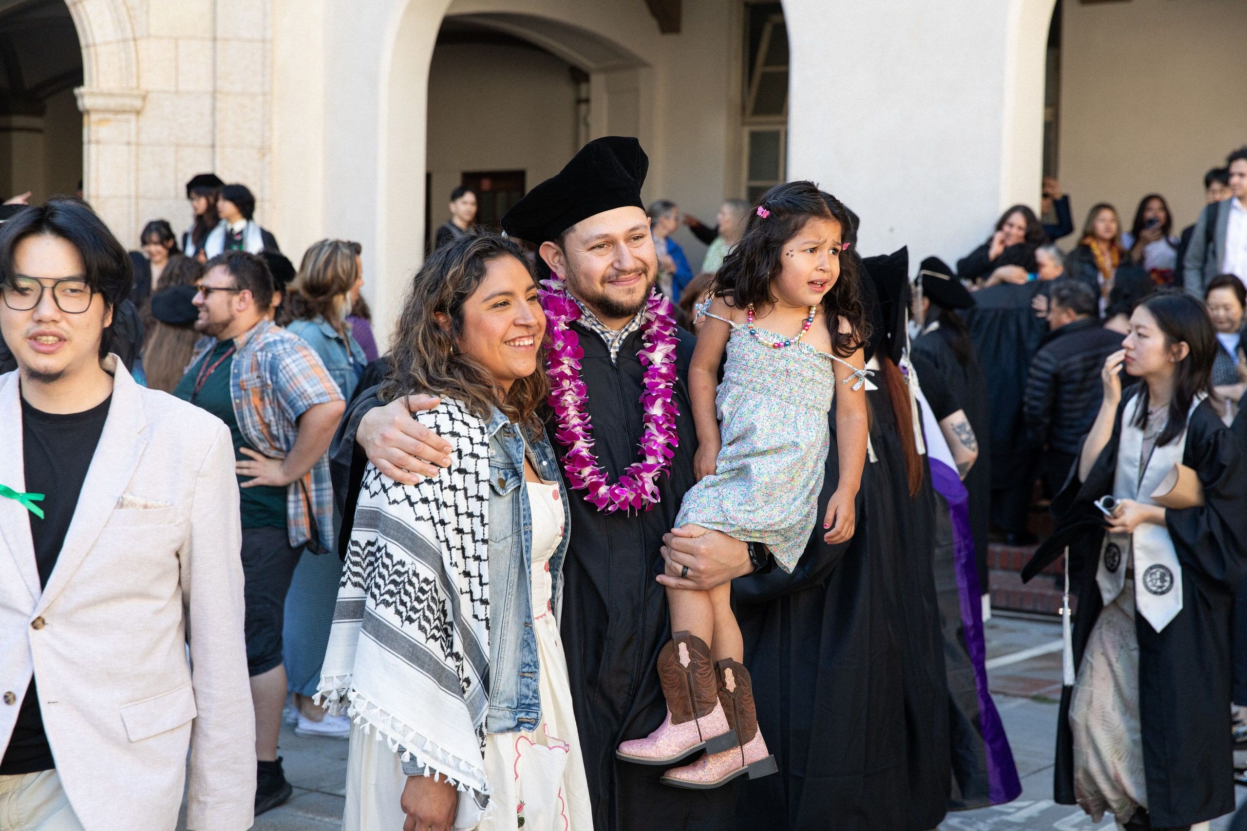 Graduates in academic regalia celebrate in a courtyard, some wearing colorful leis and sashes, with historic buildings featuring arched architecture surrounding them.