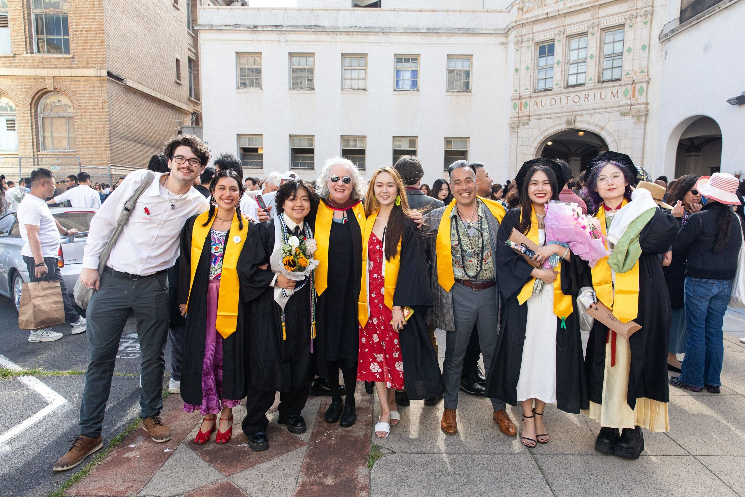 A group of graduates wearing black gowns and yellow sashes pose together in a brick courtyard, standing in front of historic buildings on the campus grounds.