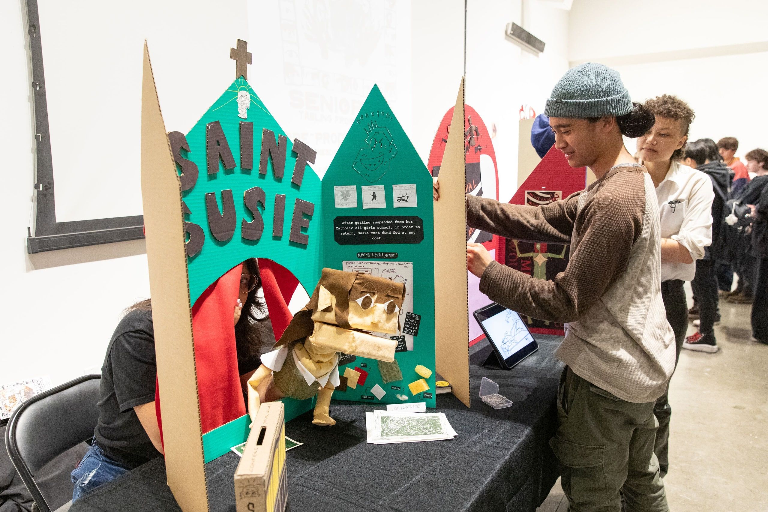 Gallery visitors interact with a colorful sculptural installation featuring turquoise and coral architectural forms with text reading 'SAINT SUSIE' at a CCA End of Year Show exhibition.
