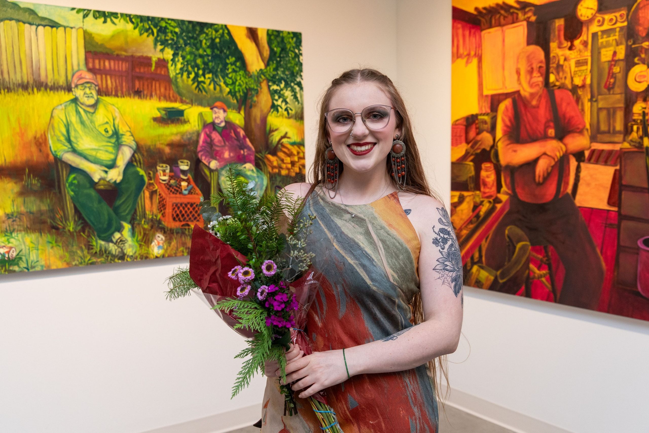 A smiling student artist holds a vibrant bouquet of flowers while standing in front of her colorful paintings featuring warm tones and figurative subjects.