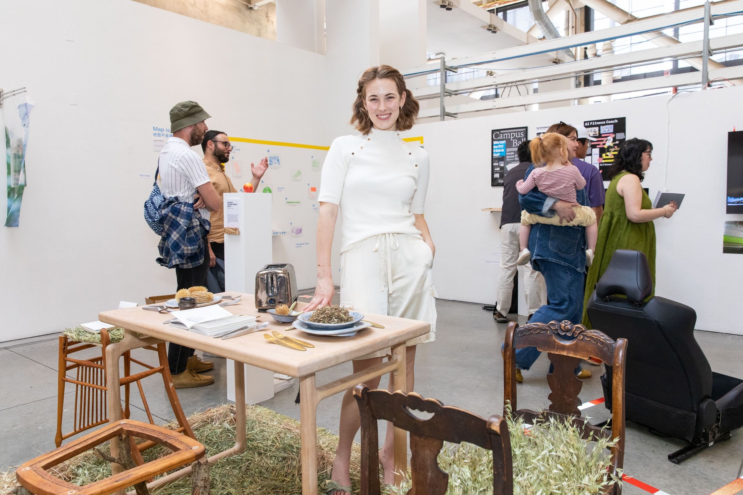 A woman in white clothing stands at a wooden table with grass-filled chair installations while gallery visitors explore the exhibition behind her.