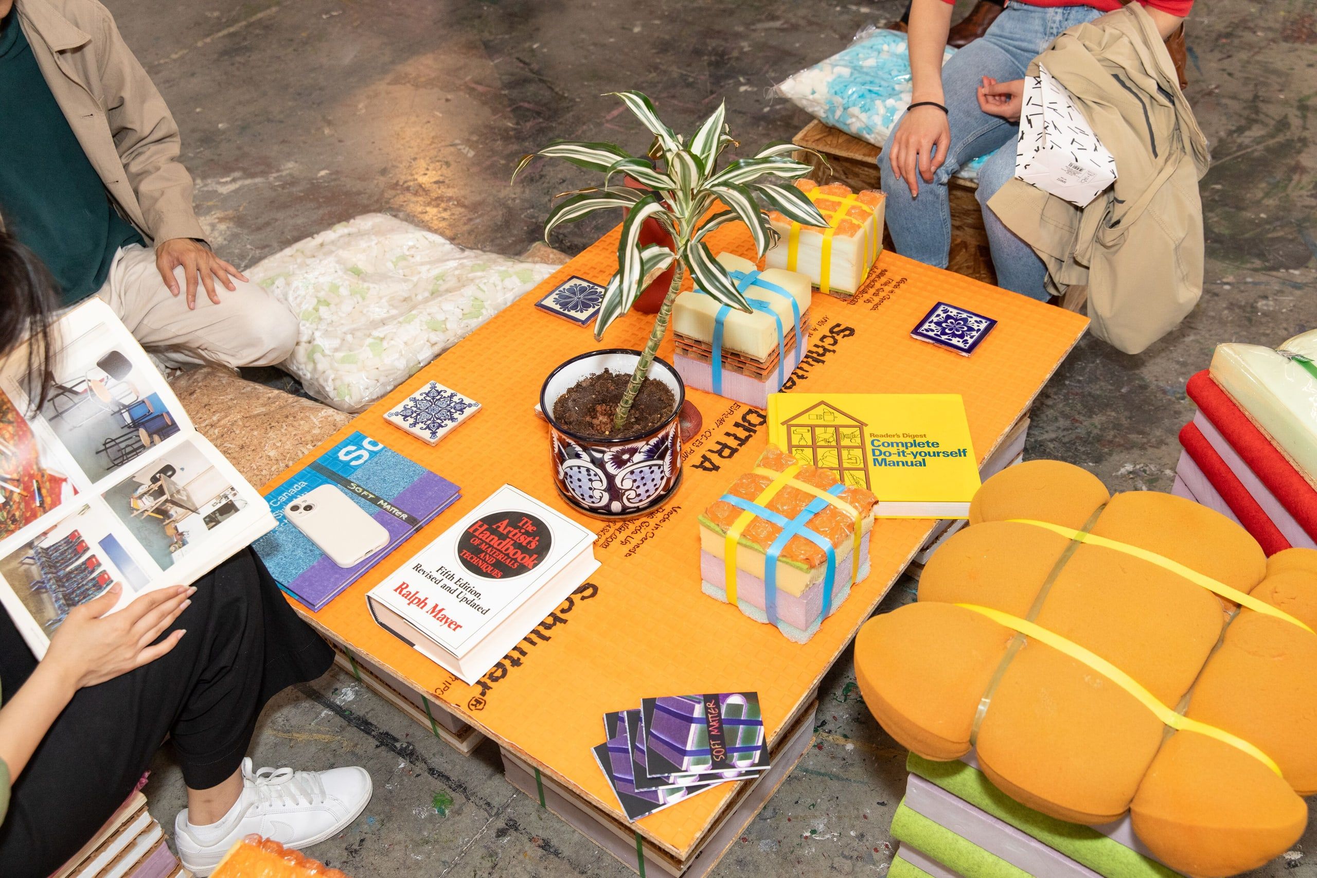 Visitors crouch around an orange exhibition display on the ground featuring a potted plant, printed materials, and colorful geometric graphics.