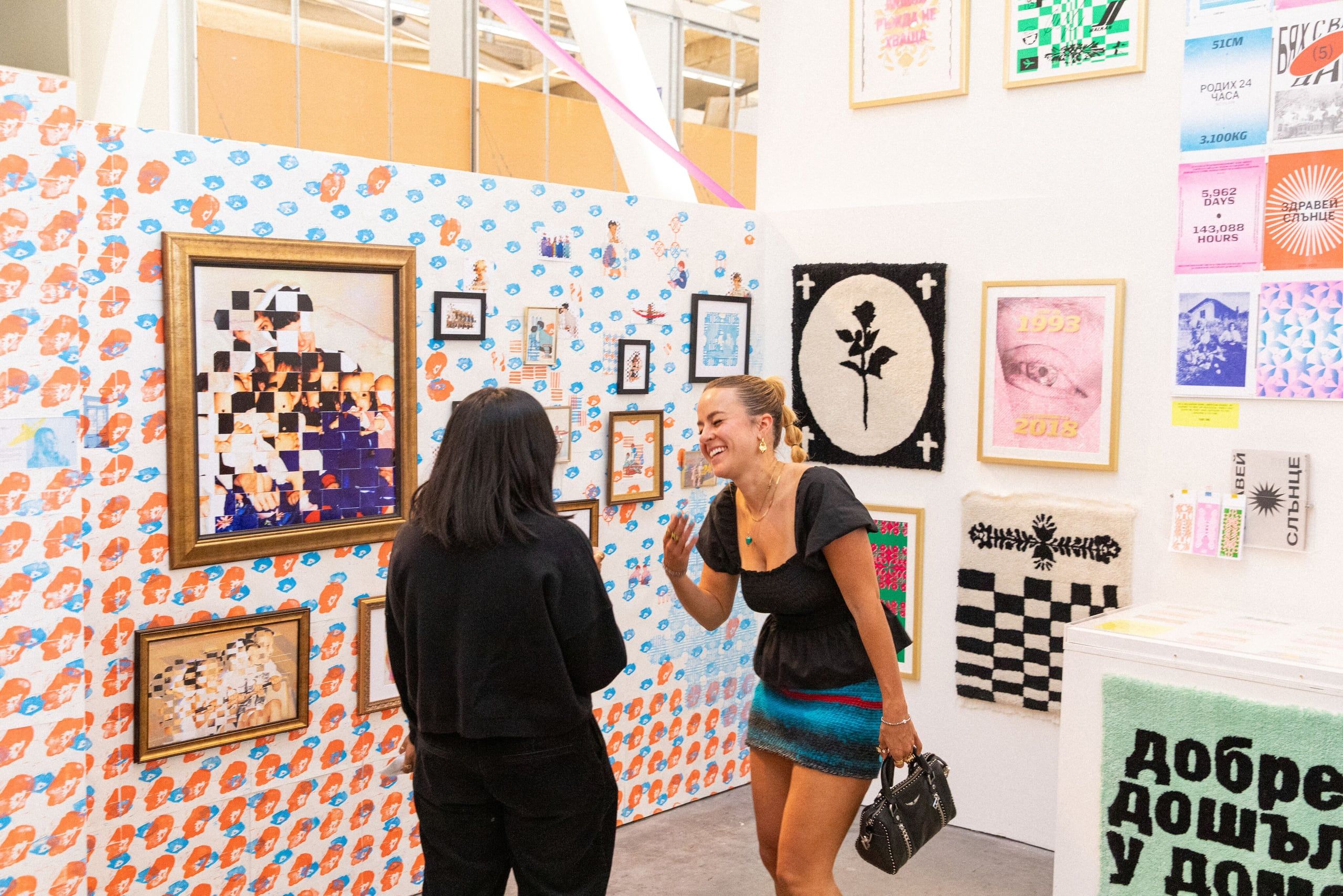 Two visitors view a gallery wall covered with colorful patterned wallpaper and displaying various framed artworks in mixed media.