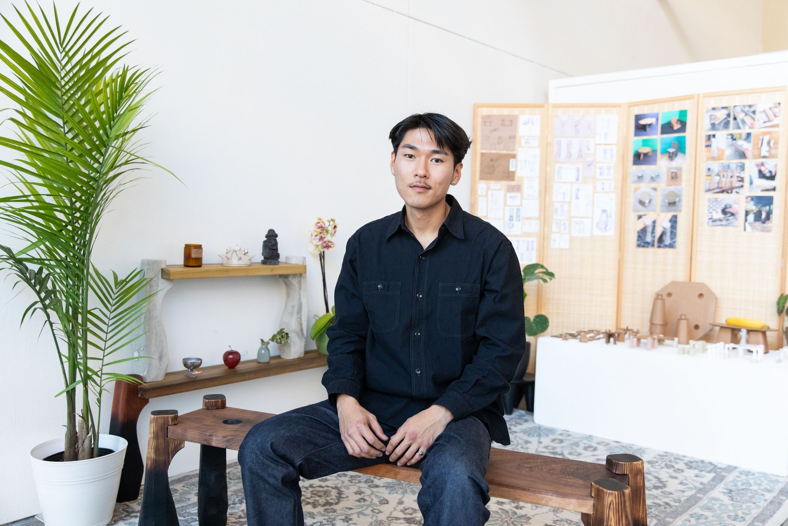 A student sits on a wooden bench in a minimalist studio space with green plants, shelving with decorative objects, and mood boards pinned to the wall behind him.