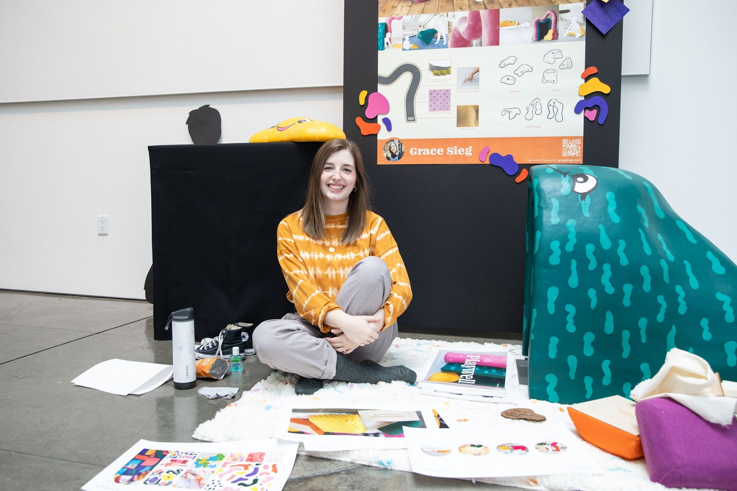 A student in a yellow patterned shirt sits cross-legged on the floor surrounded by colorful design work, sketches, and fabric swatches displayed on black panels and teal fabric.