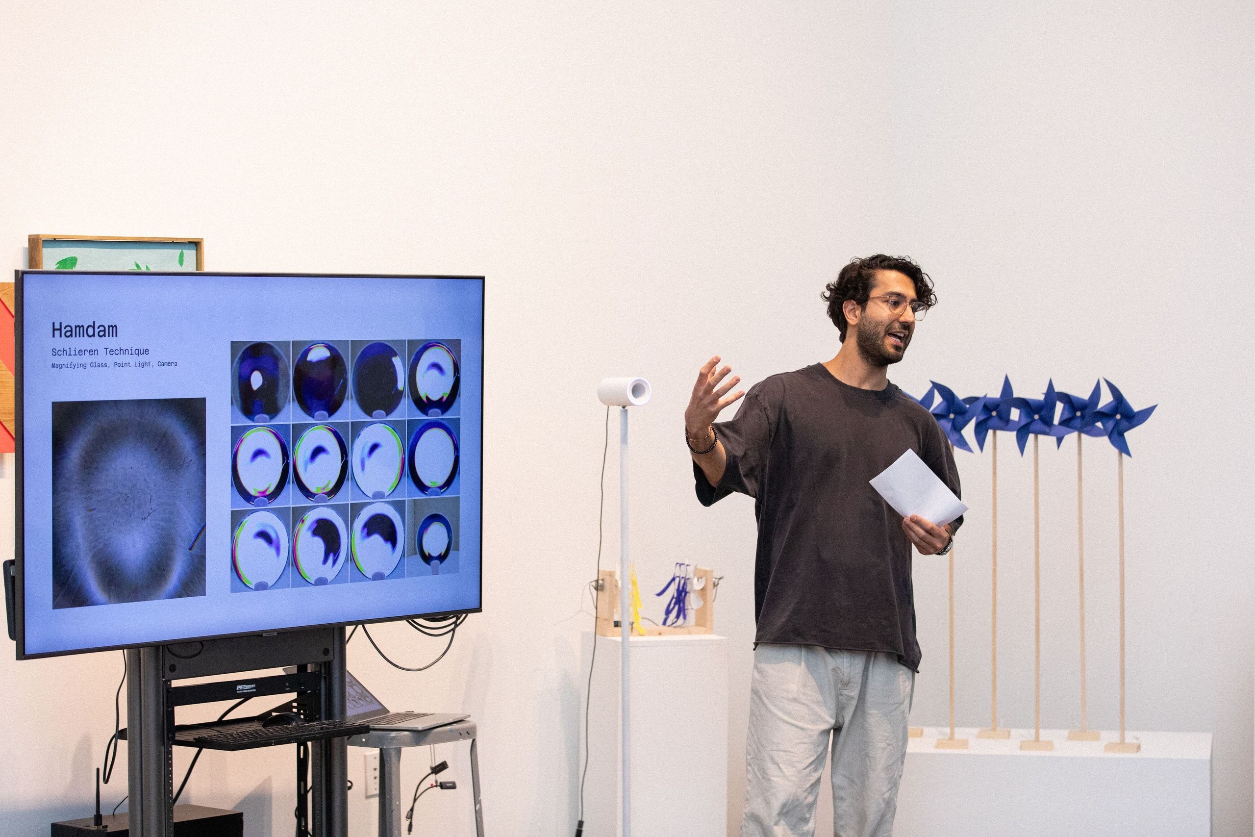 A student presents their work beside a monitor displaying brain imaging scans and a blue sculptural form with spikes mounted on the white gallery wall.