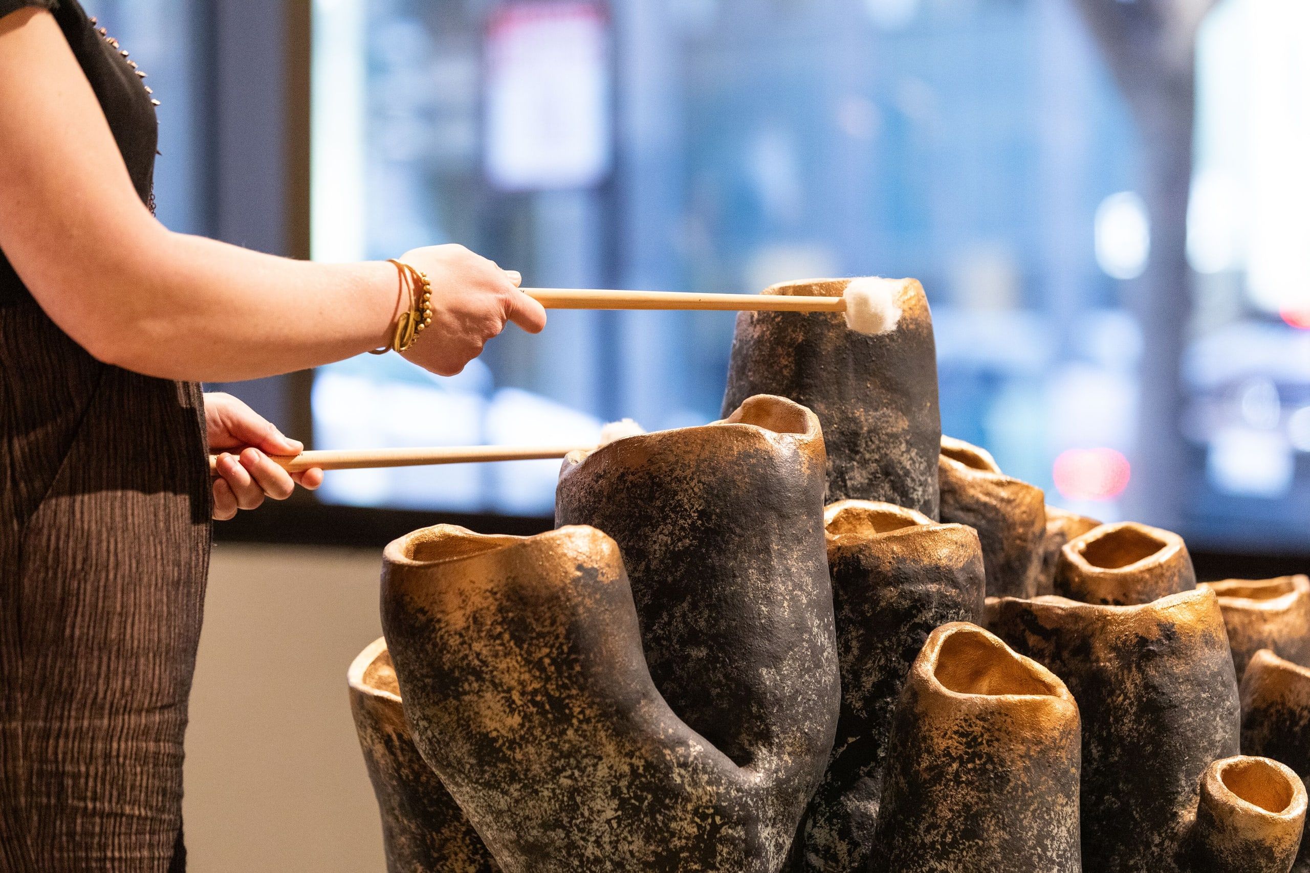 A close-up of hands using wooden tools to interact with a collection of textured, carved ceramic or wooden sculptural forms displayed at eye level.