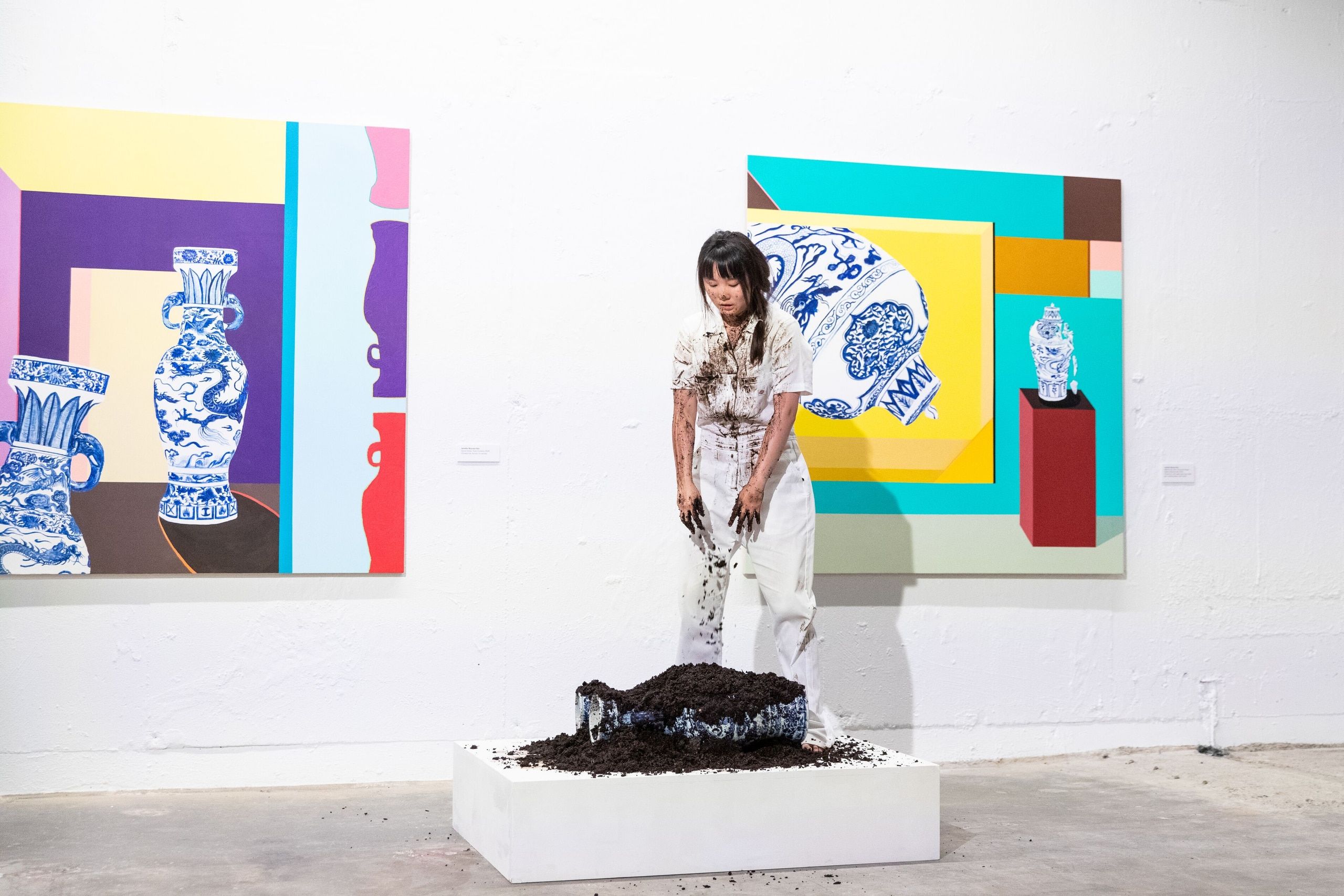A student stands before their artwork featuring colorful geometric paintings with blue and white pottery imagery and a sculptural black form emerging from the white gallery floor.